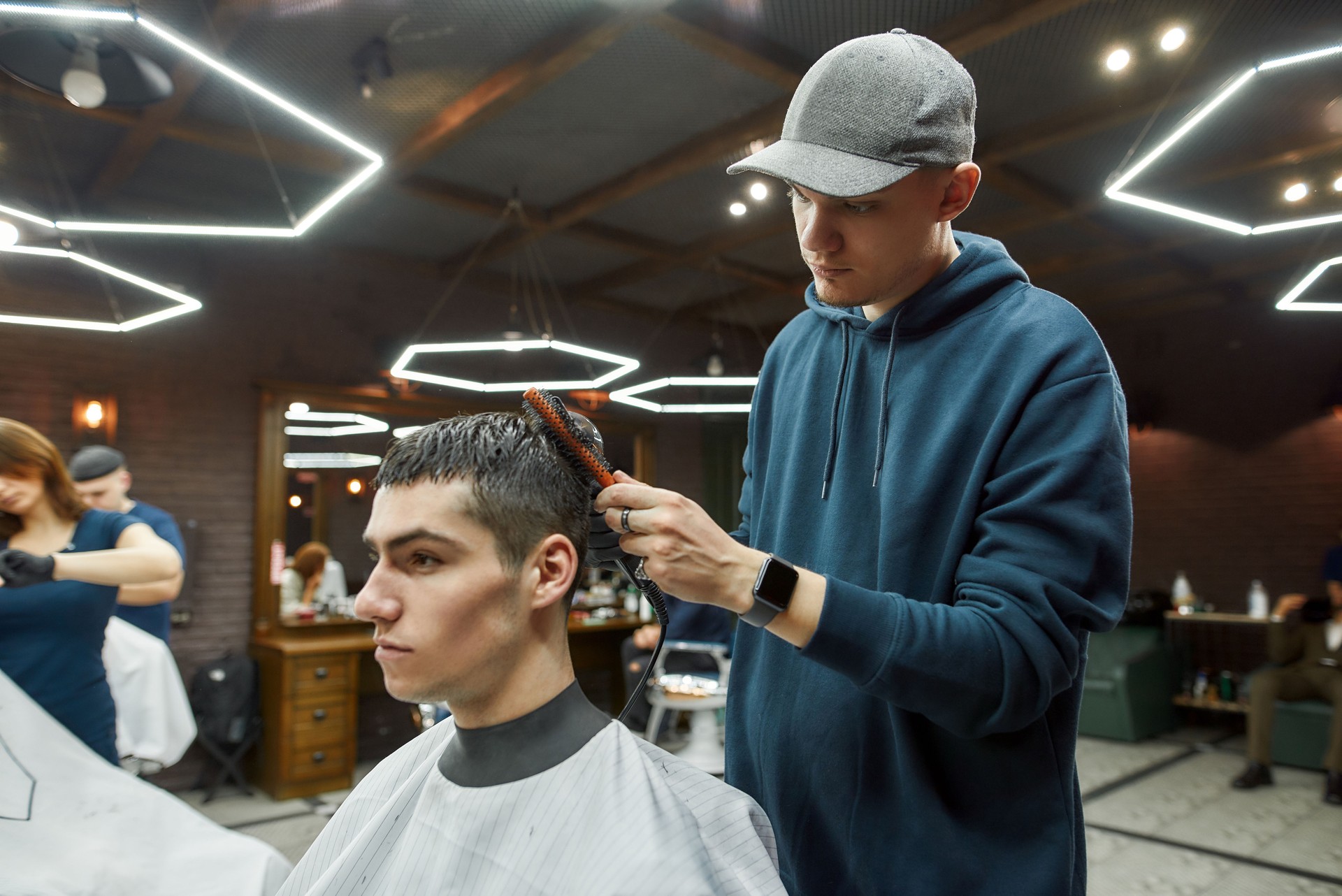 Getting trendy haircut. Side view of a professional barber drying the hair of a handsome guy in the modern barbershop, serving client